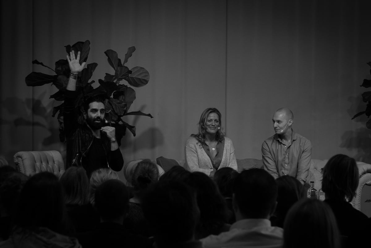 Navid Modiri står på scen med handen i luften bredvid två gäster i en soffa, publikhuvuden i förgrunden. Svartvitt.