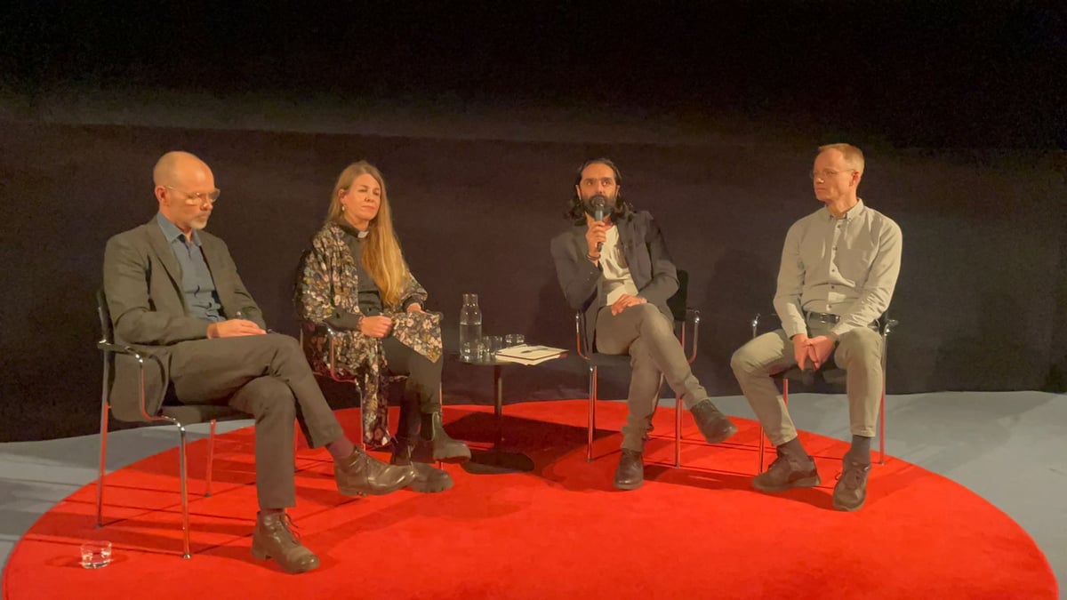 Navid Modiri talar i mikrofon under en paneldebatt med tre andra deltagare på en röd matta.